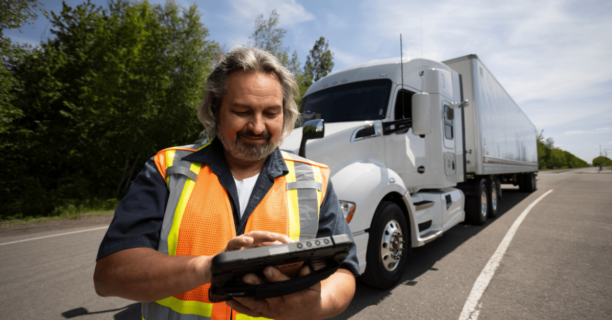 truck-driver-using-tablet-roadside Truck driver wearing a safety vest using a tablet roadside with a white semi-truck in the background.